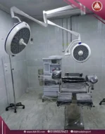 A real, wide-angle front view of a white LED 2-camera surgical light, suspended from the ceiling inside an operating room above an operating table.