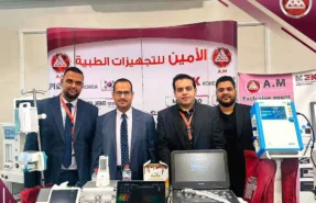 أربعة ممثلين لشركة الأمين للتجهيزات الطبية يقفون أمام جناح الشركة في معرض طبي مع أجهزة كورية.