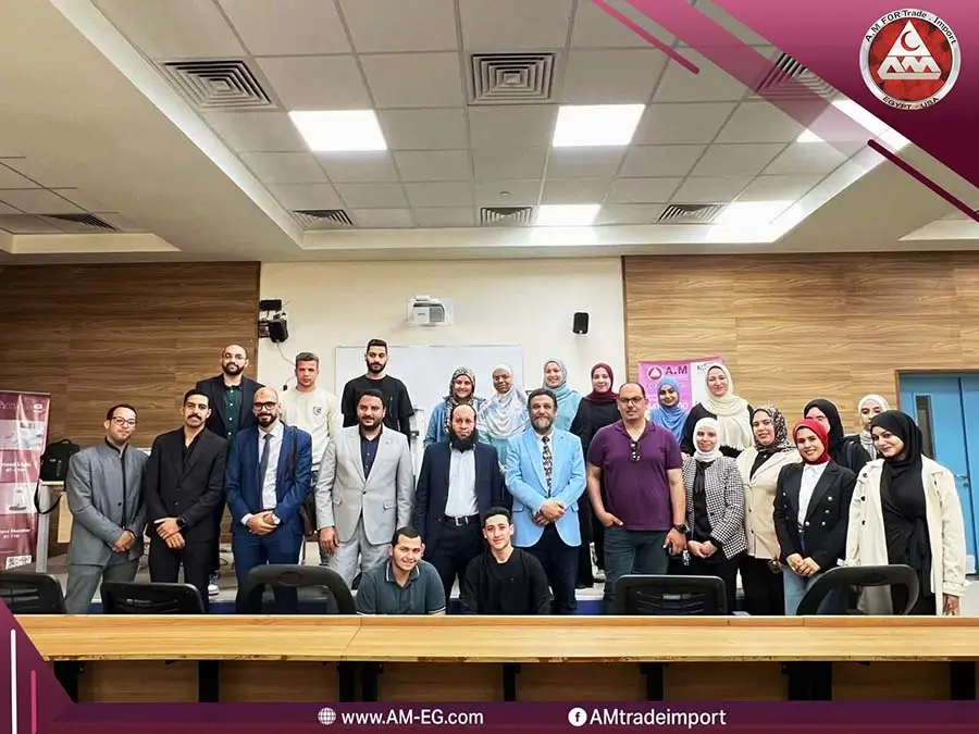 صورة جماعية لفريق شركة A.M للتجارة والاستيراد مع الحضور داخل قاعة تدريب، رجال بالبدل وسيدات بالحجاب يقفون في صفوف، وخلفهم سبورة وشاشة عرض، وشعار الشركة في الزاوية